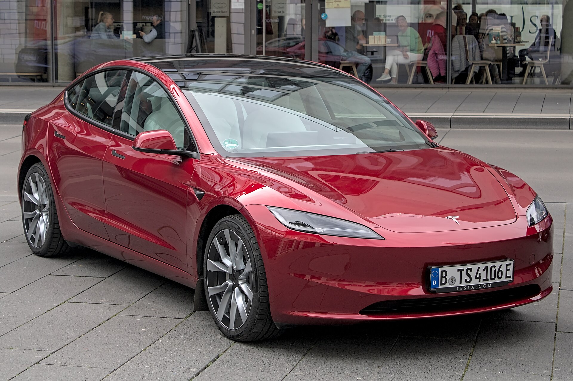Red Tesla Model 3 (Highland) shown at an outdoor auto show, front three-quarter view